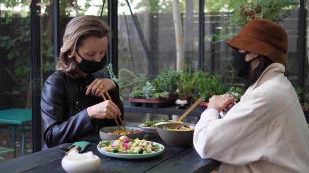 Almuerzo durante la pandemia de covid-19. Dos chicas en el restaurante vegano en máscara facial discutiendo algo sobre sus comidas veganas en estilo asiático — Vídeo de stock