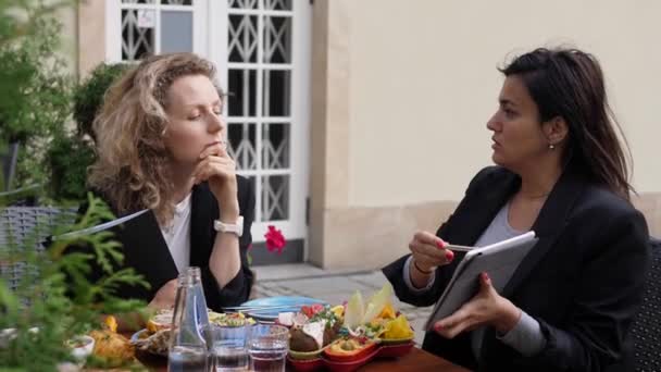 Brunette zakenvrouw toont grafieken van een ontwerp aan haar blonde collega. Zakelijke bijeenkomst tijdens de lunch — Stockvideo