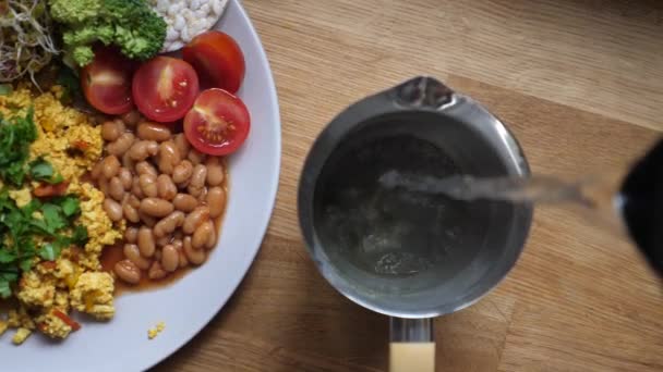 Верхній вид заливання гарячої води до чайного горщика поруч з тарілкою з повним англійським сніданком у веганському стилі — стокове відео