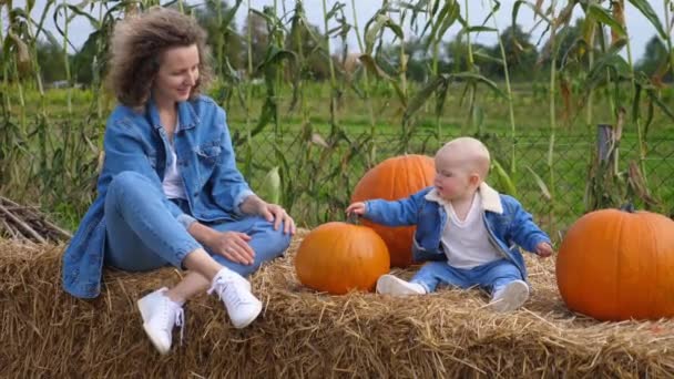 Glückliche alleinerziehende Mutter und ihr Baby im passenden Jeans-Outfit sitzen auf einem Strohballen und spielen mit den Kürbissen. Mutter-Tochter-Bindung — Stockvideo