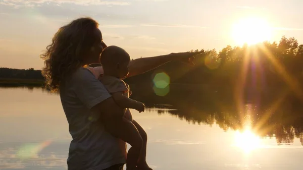 Mutlu genç anne bebeğini kucağında tutuyor ve gün batımında bir şeye işaret ediyor. Arka planda göl ve ağaçlar var. Hayatın ve doğanın tadını çıkarıyorum.. 