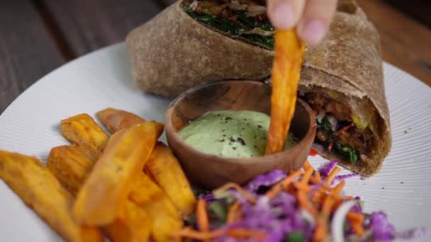 Eating clean concept. Hand dipping sweet potato in green creamy sauce. Vegan wrap and a salat served on a white plate. — Stock Video