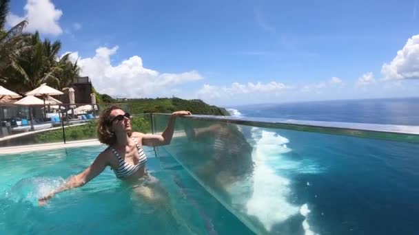 Mulher branca espirra água na piscina infinita com vista para o oceano.A câmera se move junto com o respingo — Vídeo de Stock