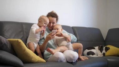 Mutlu aile kavramı. Genç anne meraklı ikizleriyle oynuyor, akıllı telefondan bir şey gösteriyor. Kanepede sevimli bir köpek var.