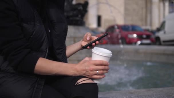 Primer plano de las manos femeninas sosteniendo el teléfono inteligente y la taza para llevar. Fuentes en el fondo — Vídeo de stock