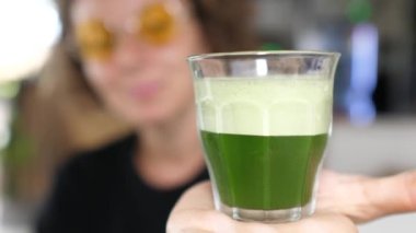 Arka plandaki bulanık kişi kameraya organik yeşil spirulinalı bir cam gösteriyor.
