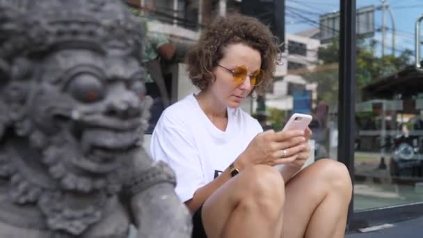 Fille caucasienne assise sur les marches à côté de la sculpture balinaise traditionnelle navigue en ligne sur son smartphone — Video