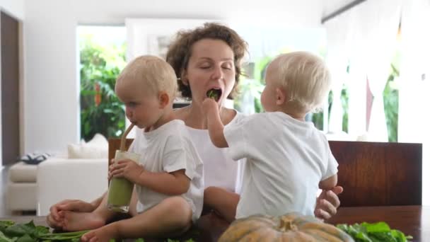 Kaukasisk baby sidder på køkkenbordet blandt grønne blade, der drikker grøn smoothie de er lige forberedt med sin unge mor. – Stock-video