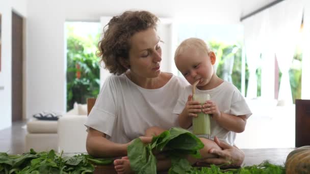 Bebé caucásico sentado en la mesa de la cocina entre hojas verdes bebiendo batido verde que acaban de preparar con su madre joven — Vídeos de Stock