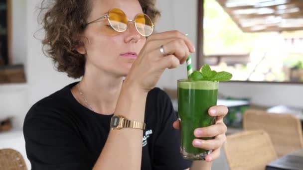 Chica caucásica con gafas de sol de color naranja revolviendo y bebiendo jugo verde fresco con paja de papel sostenible. Concepto de alimentación limpia — Vídeos de Stock