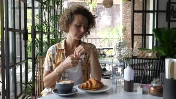 Kaukasische Frau zieht ihre Gesichtsmaske ab, um in stilvollem Café einen Kaffee mit Croissant zu trinken. Leben während der Coronavirus-Pandemie — Stockvideo