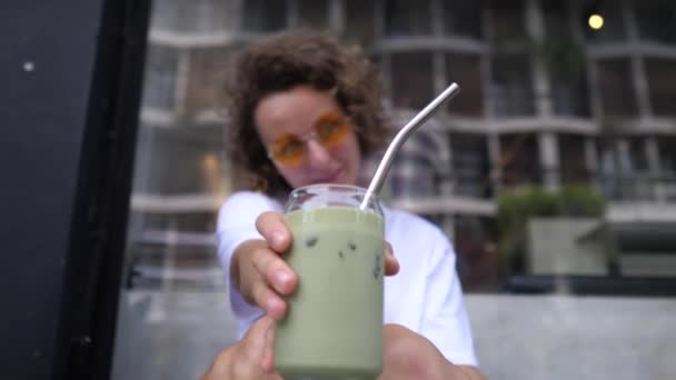 Glada blondin visar sin ekologiska växt baserad matcha is te latte i en glasburk med metallhalm till kameran — Stockvideo
