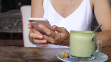 Kahve dükkanında bir kadının mesajlaşmasını kapat. Bitki bazlı kibrit sütlü latte, yanında kurabiye olan bir masada servis edilir.