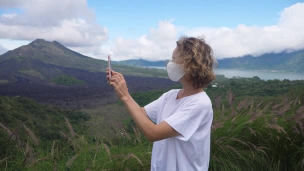 Vista lateral de la joven mujer caucásica en una máscara facial tomando fotos de las montañas con su teléfono inteligente. Viajar solo durante la pandemia de covid-19 — Vídeo de stock
