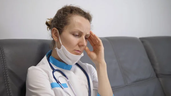 Frustrated Caucasian doctor lady takes of her face mask and rubs her temples