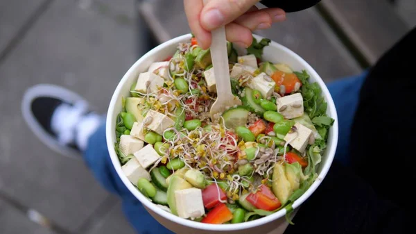 Healthy food delivery for quick meal on the go. Close up of hand mixing green veggie salad with sprouts, tofu, microgreens in recyclable paper pack