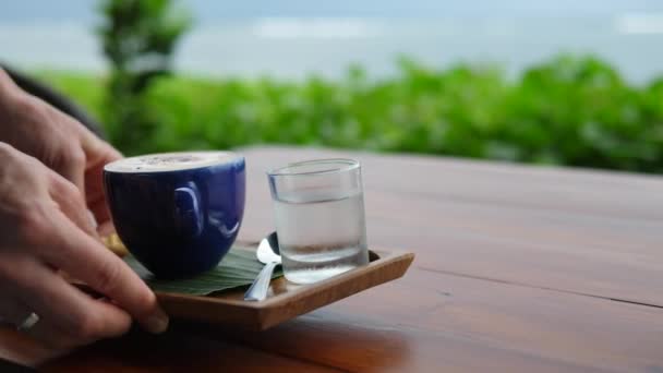 Manos sirviendo macchiato expreso con galleta de cortesía en bandeja de madera. Pausa para café con vista al mar — Vídeos de Stock