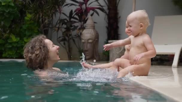 Concepto familiar feliz. Bebés gemelos caucásicos sentados en el borde del agua salpicada de la piscina. Mamá dentro de la piscina — Vídeos de Stock