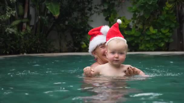 Yüzme havuzunda Noel Baba şapkası takan mutlu büyükanne ve bebek. Tatiller ve Tatil Konsepti. — Stok video