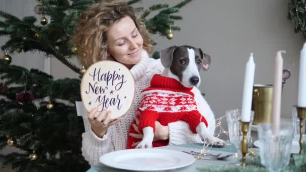 Cena para dos mejores amigos. Chica caucásica mostrando Feliz Año Nuevo signo y linda mascota en ropa de abrigo en la mesa puesta en la decoración festiva — Vídeos de Stock