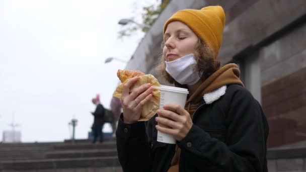 Veřejné pouliční obchody s potravinami uprostřed omezení covid-19. Mladá žena odstraňuje obličejovou masku k pití matcha latte s čerstvě upečeným croissantem — Stock video