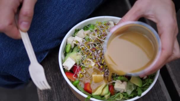 Ovanifrån av hand dressing rå vegan sallad med tahini olja i endosbehållare. Friska mellanmål för lunchrast utomhus — Stockvideo