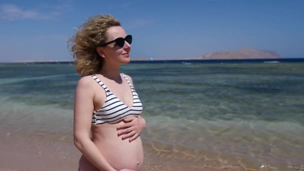 Férias à beira-mar para pais recém-nascidos. Mulher feliz em maiô com barriga grávida passeia ao longo da praia — Vídeo de Stock