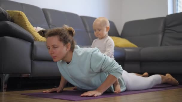 Funny kid sitting on moms back during her home yoga class. In-house physical activity and sport together with kids — Stock Video