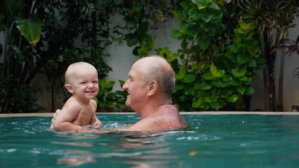 Grandfather playing in the swimming pool with his grandchild. Happy family on holidays