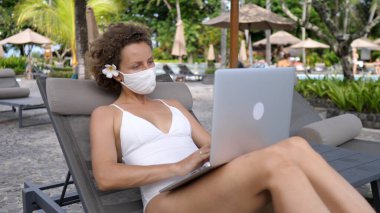 Salgın sırasında kumsalda uzaktan çalışma. Koruyucu maskeli genç beyaz kadın havuz başında güneşlenirken dizüstü bilgisayarla çalışıyor.. 