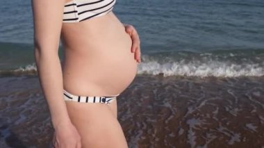 Bikinili hamile bir kadın sahilde karnının üzerinde yürüdüğünü hissediyor ve yakında bir çocuk bekliyor. Yaz tatilleri için deniz tatil beldesi