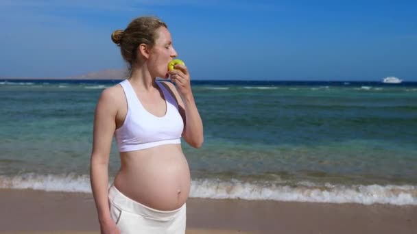 Gezonde prenatale voeding tijdens gezinsvakanties. Sportieve zwangere vrouw eten appel door de zee genieten van een gelukkige levensstijl — Stockvideo