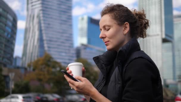 Vida en ciudades metropolitanas. Mujer casual en descanso trabajando desde el teléfono con llevar café en una zona rica con edificios de gran altura — Vídeos de Stock
