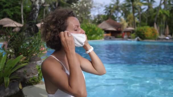 Jonge blanke vrouw draagt een beschermend gezichtsmasker tijdens het chillen bij het zwembad. Reizen tijdens een corona-viruspandemie — Stockvideo