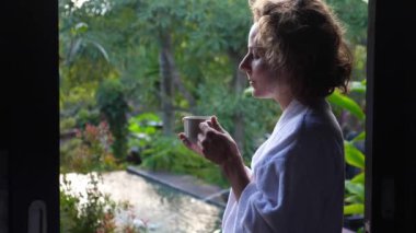 Mükemmel sabah ritüelleri. Kadın sıcak kahveden bir yudum alır. Balkonda durur ve tropik bahçenin tadını çıkarır.