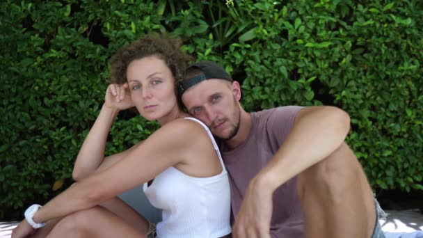 Portrait of young white couple sitting in front of leafy wall. Man leaning on woman shoulder — Stock Video