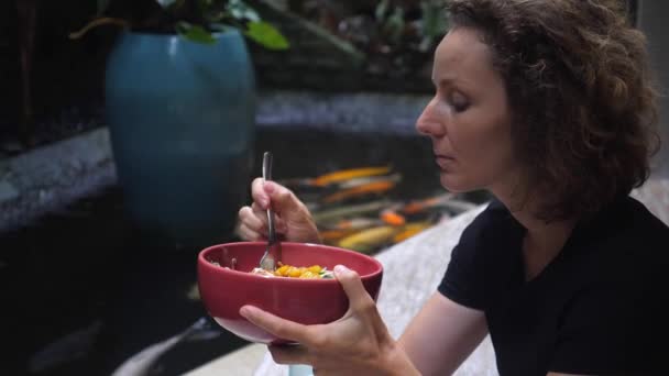 Chica caucásica come su vegano saludable megan en el restaurante con un estanque con carpas koi en ella. REstaurant con el concepto de vistas — Vídeos de Stock