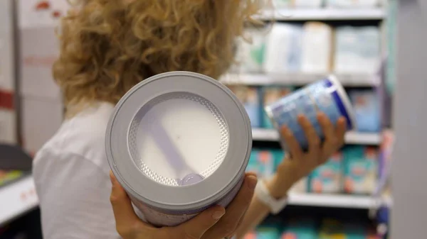 Maskeli bir kadın bebek formüllerini karşılaştırıyor, etiketleri okuyor ve birini seçiyor. Bebek maması konservesi tutarken yakın plan. COVID-19 salgını sırasında temel alışveriş.