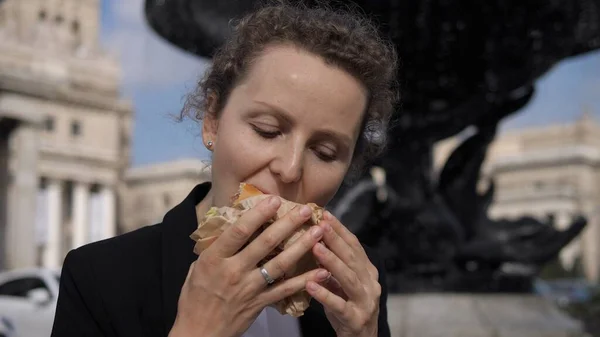 Hungrig kvinnlig kontorsarbetare äter hamburgare vid hennes lunch utomhus nära fontänen — Stockfoto