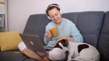 Dizüstü bilgisayara bağlı kulaklıklarla kanepeye kilitlenmiş, köpeği olan serbest çalışan bir kadının yan görüntüsü. Covid-19 kendini izole etme kavramı. 