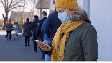 Kadın süpermarkete girmek için kuyrukta beklerken akıllı telefon kullanarak yüz maskesi takıyor. Coronavirus salgını. Bialystok, Polonya - Mart 2020.