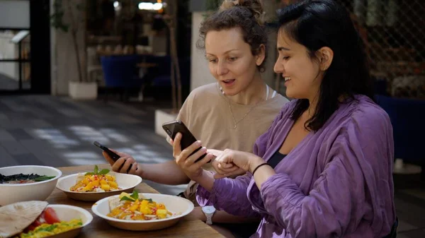 Deux copines joyeuses se montrant quelque chose de drôle dans leur téléphone pendant le déjeuner. Profiter d'une délicieuse nourriture saine et du temps de qualité passé avec des amis. — Photo