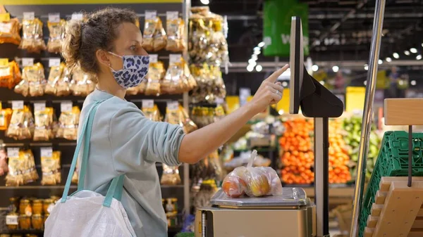 Vue latérale d'une fille blonde dans un masque facial pesant des fruits sur une balance électronique dans un supermarché. Shopping pendant la pandémie de covide-19 — Photo