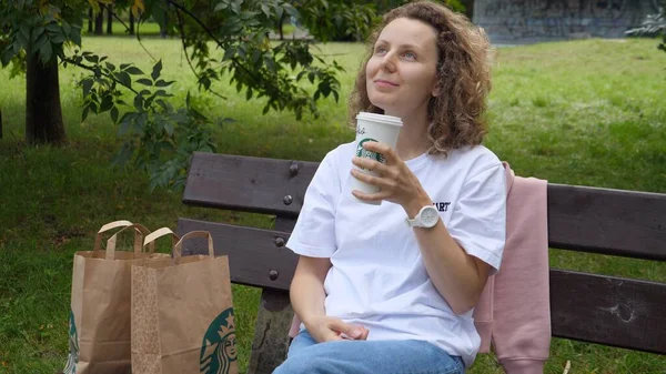 Белая девушка в парке сидит на скамейке и наслаждается напитком Старбакс. Warsaw-Poland-2020 — стоковое фото