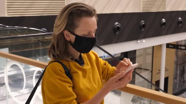 Compras durante a pandemia do covid-19. Mulher jovem mensagens de texto enquanto espera por alguém usando máscara facial para evitar a propagação do vírus corona — Vídeo de Stock