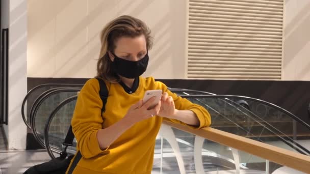 Einsame Frau mit Gesichtsmaske steht mit ihrem Handy im Einkaufszentrum neben Rolltreppen — Stockvideo