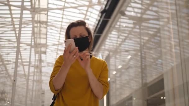 Busy cheerful girl with protective face mask walks looking at her phone in in shopping center. Glass roof over her head — Stock Video