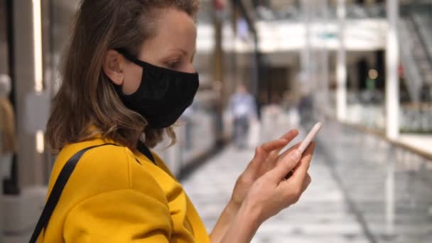 Vedere laterală a femeii cu mască de față folosind smartphone-ul ei în mall în timpul pandemiei covid-19 — Videoclip de stoc