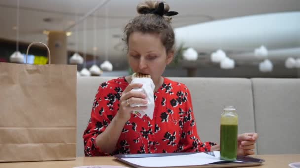 Jovem com um pão de cabelo comendo um sanduíche e bebendo smoothie verde na praça de alimentação do shopping — Vídeo de Stock