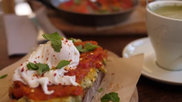 Gezonde vegetarische mediterrane brunch uit. Avocado toast met gepocheerd ei en tomatenpasta op houten bord met koffie — Stockvideo